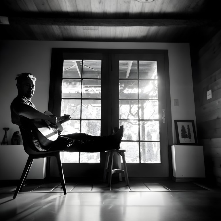 Black and white image showcasing Michellion playing guitar in front of a large glass door, where sunlight creates an interplay of shadows and highlights, symbolizing the contrasts in life and music.
